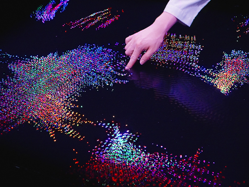 Living Crystallised Light (2025), from the series Living Crystallised Light (2022), invites visitors to dip a finger into a glimmering pool, where ripples bloom into a dynamic mosaic of light. (Image Credits: Courtesy of teamLab and Pace Gallery)