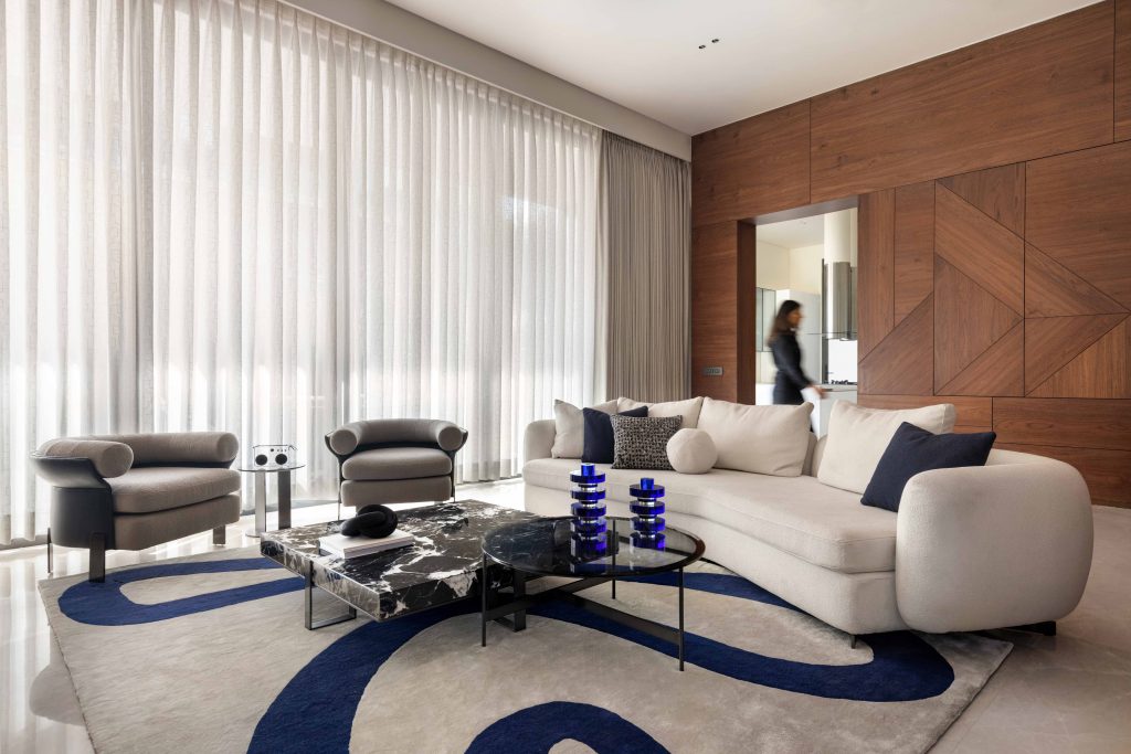 A sculptural sofa inspired by a Poliform design is accompanied by tactile armchairs and a fluid-patterned rug by Prince Home Furnishing with deep blue accents, adding quiet luxury to the informal living room. (Image Credits: Purnesh Dev Nikhanj)