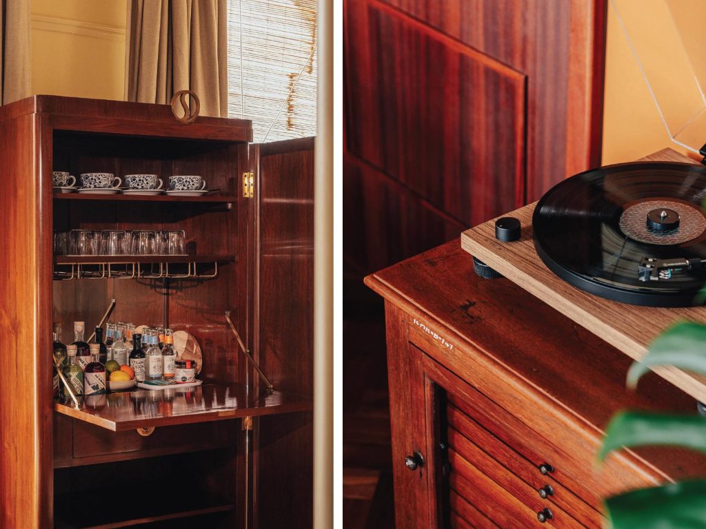The Art Deco bar, accented with gleaming brass details, and a seasoned teakwood turntable console with fluted shutters, anchors the space in nostalgic elegance.Image Credit: (Permit Room Lodgings)