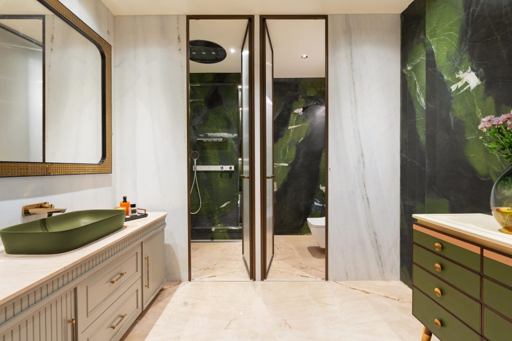 The bathroom is wrapped in striking floor-to-ceiling Brazilian Avocado Quartzite stone, its rich veining bringing depth and natural drama to the space. A statement mirror from Apartment 9 adds a touch of sculptural elegance, while vases from Armoire offer a refined, organic accent that softens the bold material palette. (Image Credits: Gitesh Gupta, Studio Bluora)