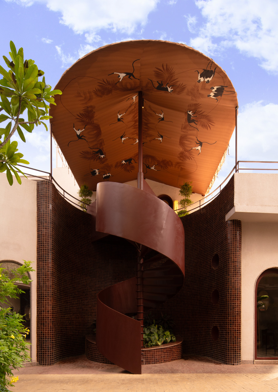 The spiral staircase unfurls like a helical sculpture, its oxblood finish cutting a bold silhouette against the mosaic-tiled courtyard wall. (Image Credits: Avesh Gaur)