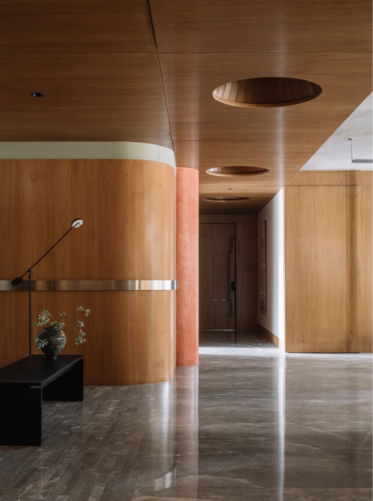 The entrance foyer is anchored by sleepy grey marble and warm wooden panelling with a ceiling that features recessed circular up-throw lights that cast a glow onto polished stainless steel discs, simulating the soft glow of a skylight. The lighting is by Smart Light. (Image Credit: Ishita Sitwala)