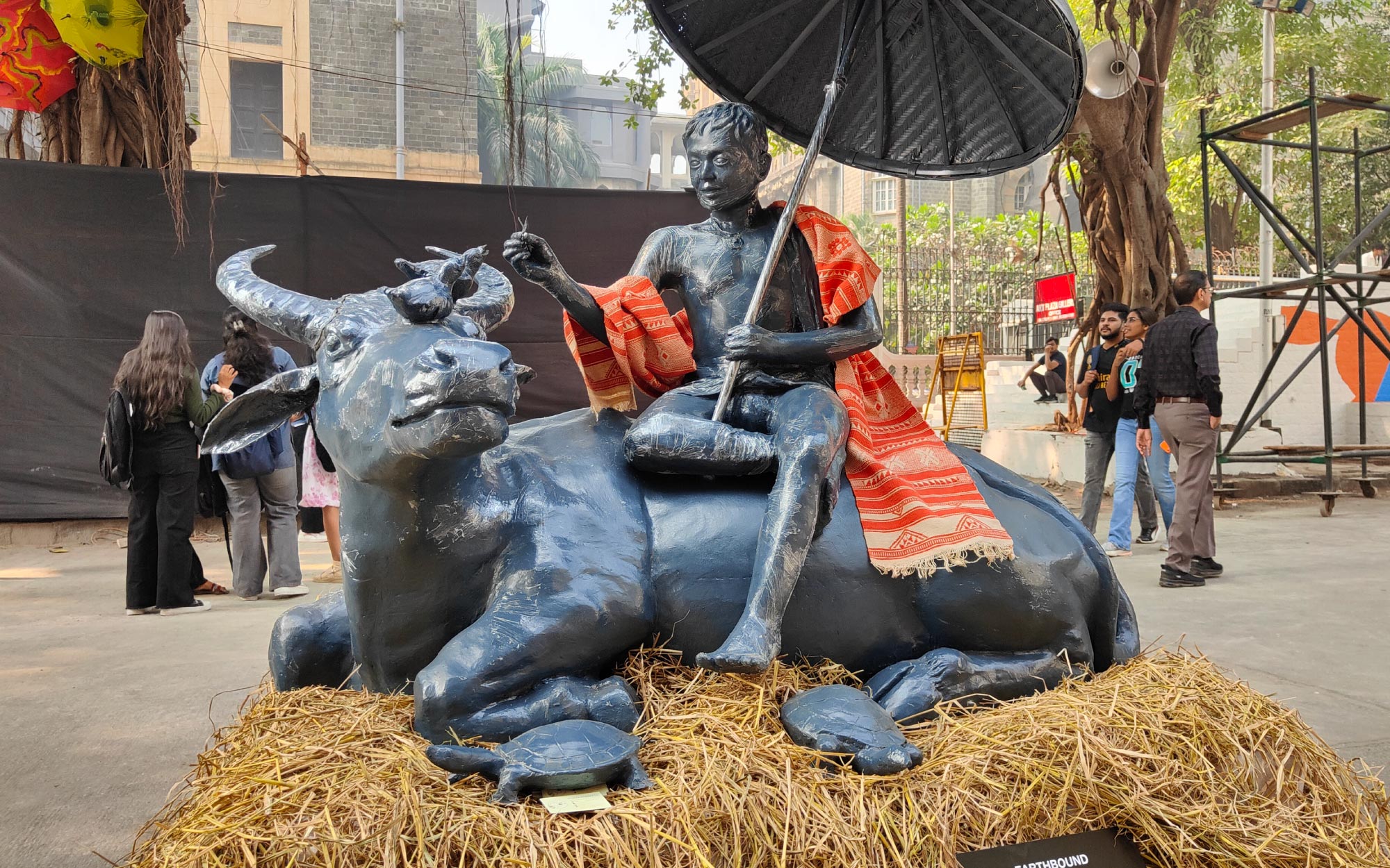 At KGAF 2024, Kerketta Art Studio presents Earthbound, depicting a tribal shepherd in repose, feeding mynahs atop his buffalo as a tortoise rests nearby. (Image Credits: Brinda Miller)