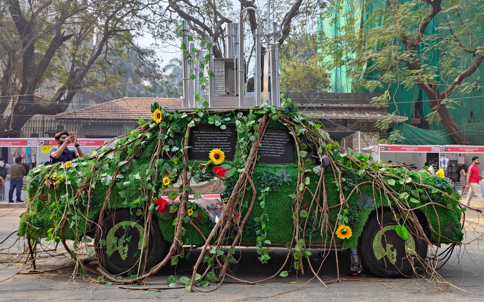 ‘Plastic Plants’ at Kala Ghoda 2024 explores environmental pollution through a car wrapped in artificial greenery, with exhaust pipes rising above to reflect the toxic overlap between manufactured nature and industrial ‘plants.’ (Image Credits: Brinda Miller)