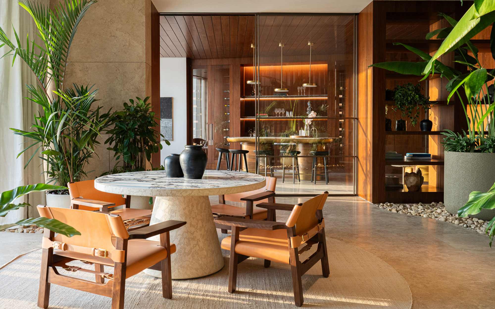 A custom marble top table and leather-wrapped chairs sit amidst an indoor garden with oversized planters in an airy glass enclosure near the living area. (Image Credit: Patricia Goijens)