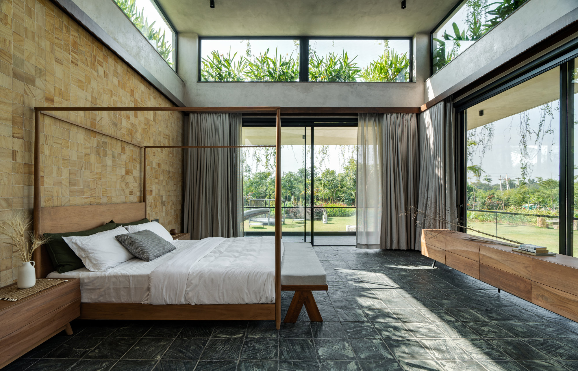 An imposing four-poster bed by Dbodhi & Otazen boasts an avant-garde ottoman at its foot. A long console in exposed live-edge wood frames the bottom half of the front window. Pale grey curtains by Skipper Furnishings match the cool Markapur slate floor. (Image Credits: Shamanth Patil)
