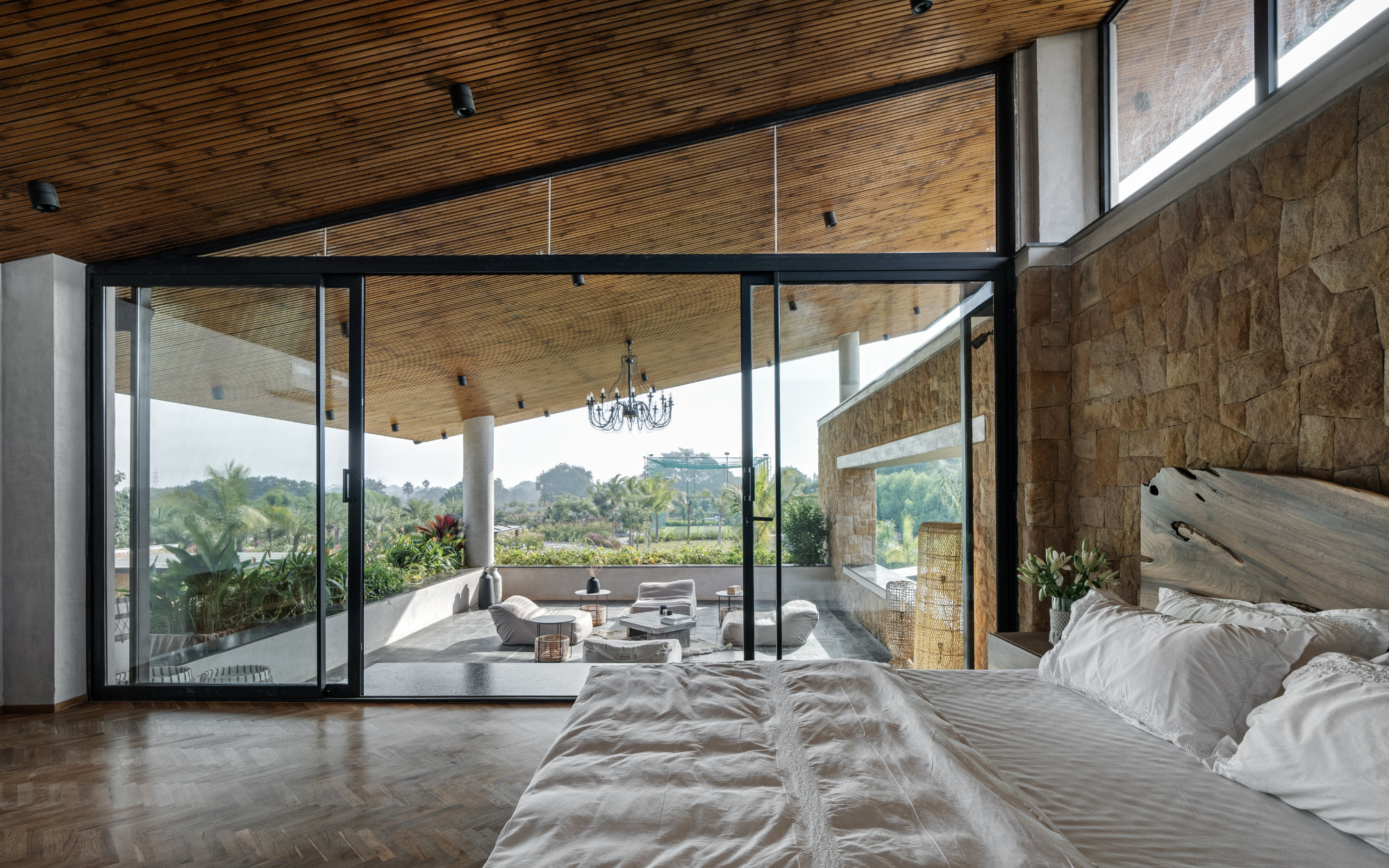 In the only bedroom placed on the first floor, Chevron-patterned parquet floors lie solemnly in contrast to the black metal accents of the glazing by Pinkmondo. A lengthy wall of Khamman brown sandstone extends beyond the bedroom onto the covered sit-out. (Image Credits: Shamanth Patil)

