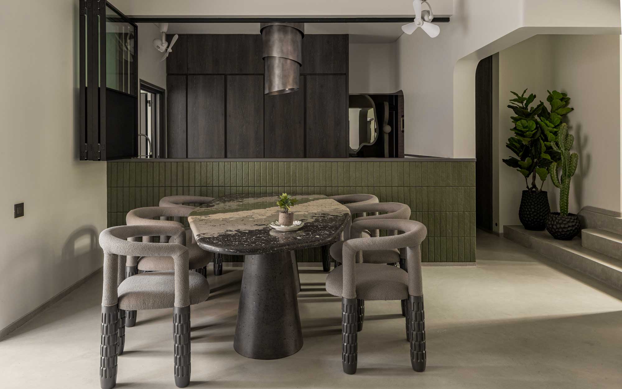 The modular kitchen by Shambhala echoes the green hues of the dining table top. A retractable chimney in burnished copper by Faber, India, marks a visual centrepoint. Sliding-folding shutters by Veneto maintain a seamless connection with the dining area. (Image Credits: Vivekajeet)
