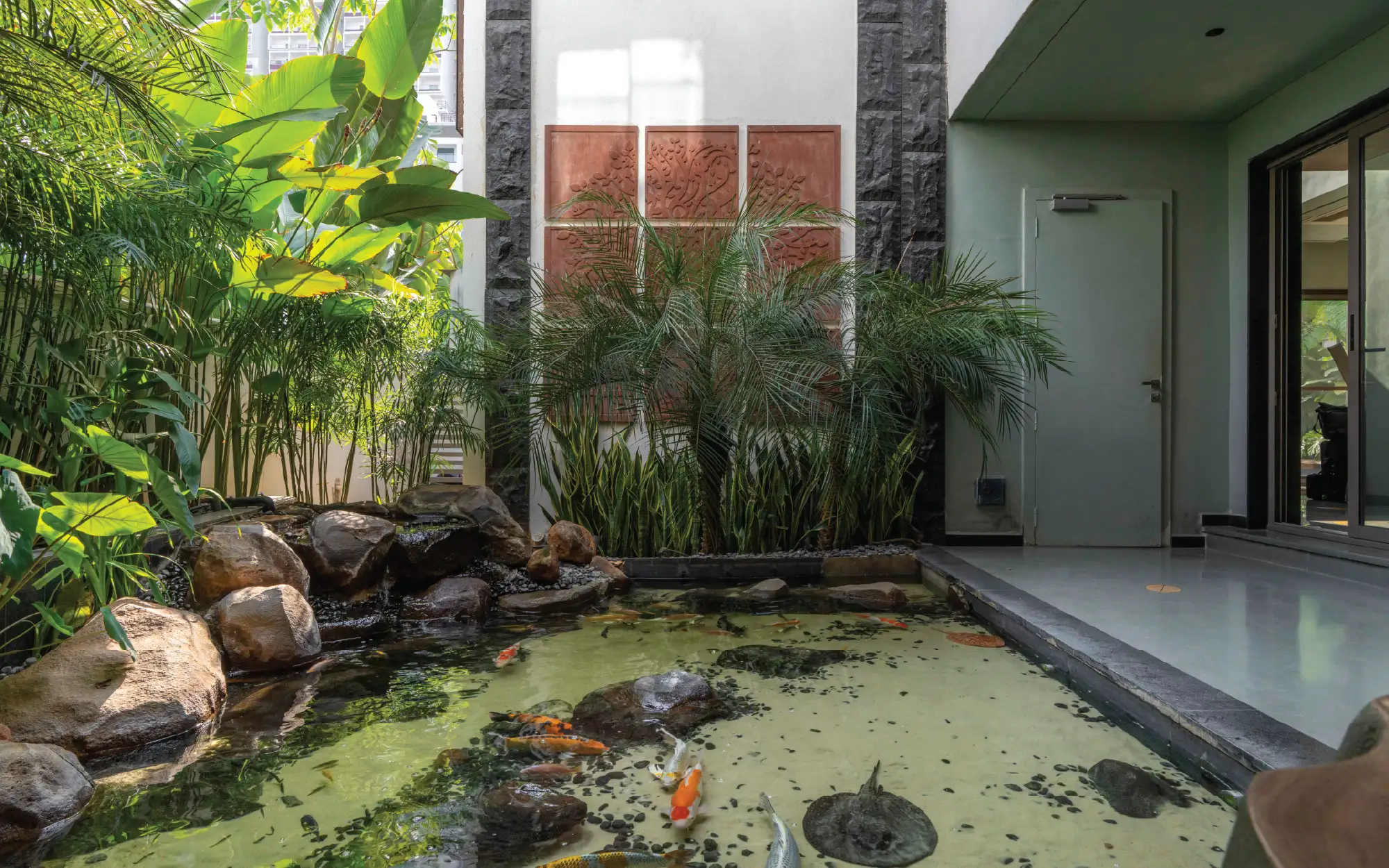 A courtyard with a tranquil koi pond, where natural boulders, antique mud mural tile frames, and lush tropical planting create an immersive landscape inside a home. (Image credits: Ricken Desai)